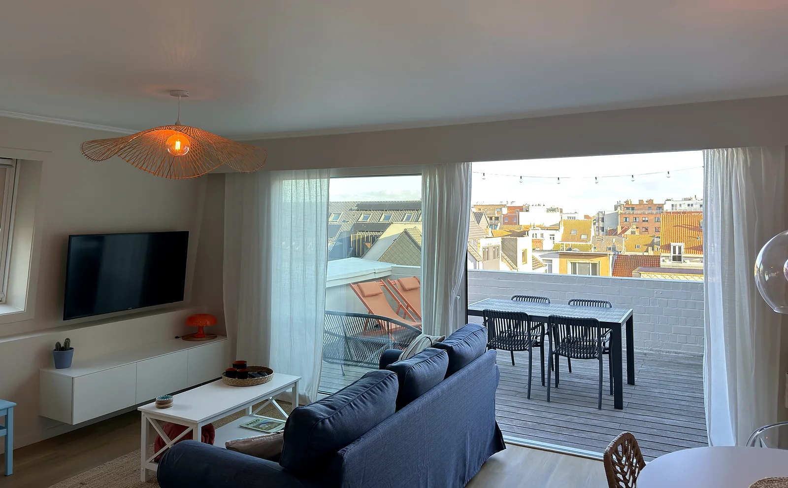 Séjour lumineux avec canapé bleu, télévision et baie vitrée ouverte sur la terrasse et les toits